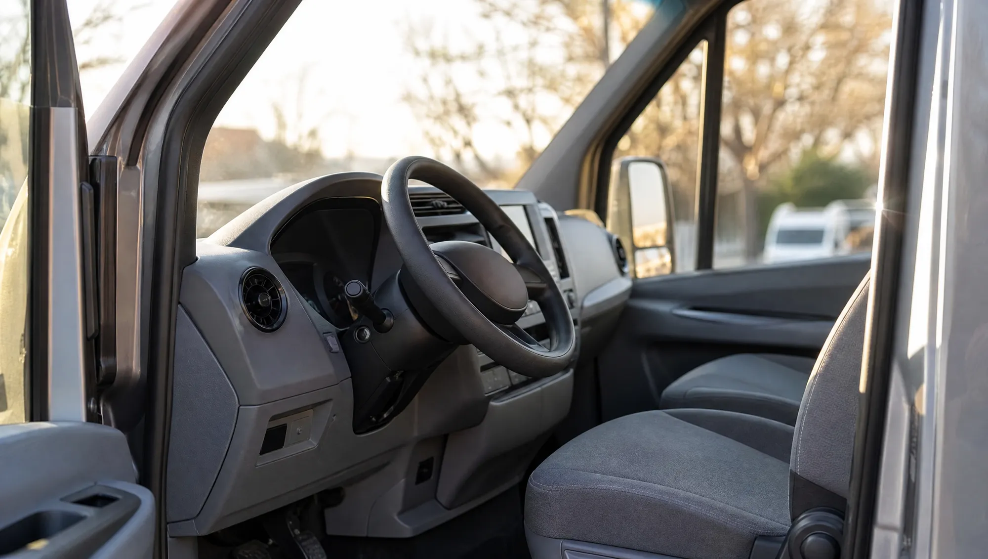 The driver's seat and steering wheel of a minibus seen through the open cab door in morning light